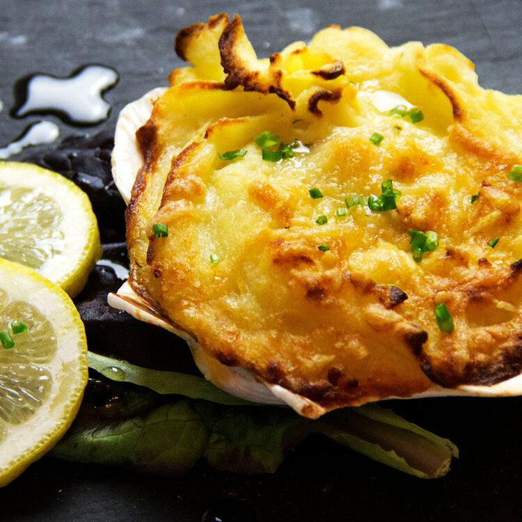 A scallop shell filled with a creamy seafood dish, topped with potatoes and cheddar cheese, served with lemon slices on the side.  Coquille St Jacques